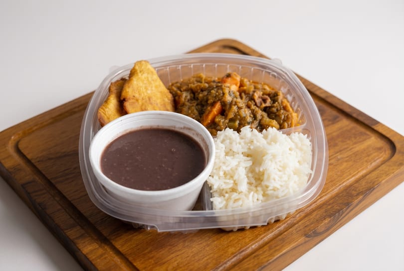 A takeout container with rice, black beans, stew, and fried plantains on a wooden board.